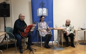 Dw&oacute;ch mężczyzn siedzi na krzesłach, jeden z gitarą, pomiędzy nimi kobieta