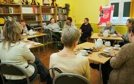 Grupa ludzi siedzi przy kilku stolikach, w tle regały z książkami. 