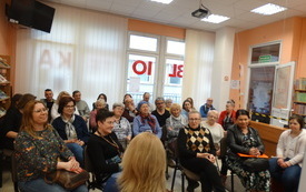 Uczestnicy spotkania, kobiety i mężczyźni. W tle okno z napisem biblioteka. 