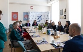 Bibliotekarze siedzący przy stole. Obok stoi Dyrektor biblioteki w zielonej marynarce i spodniach. 