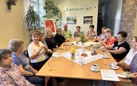 Grupa ludzi siedzi przy stole i rozmawia. w tle roślina, okno i plakat Dyskusyjnych Klub&oacute;w Książki.