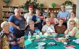 Część ludzi siedzi na krzesłach, a część stoi, trzymają w rękach logo DKK.