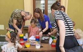 Dziedziniec biblioteki. Dzieci i dorośli układają rolki taśmy na stole. 