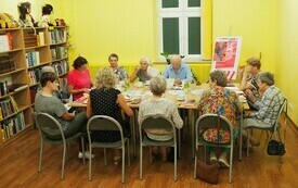 Grupa ludzi siedzi dookoła stołu, po prawej stronie logo Dyskusyjnych Klub&oacute;w Książki. Po lewej stronie regały z książkami. Na regale pluszowe maskotki misi&oacute;w. 