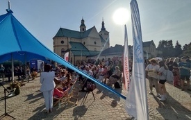 Grupa starszych i młodszych os&oacute;b siedzi na krzesłach i leżakach na wybrukowanym placu, w tle za nimi budynek kościoła