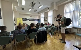 Sala konferencyjna. Liczna widownia. Mężczyzna z kamerą, filmujący spotkanie.