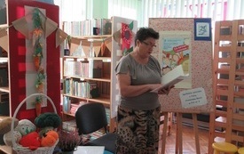 Wnętrze biblioteki. Regały z książkami, stoliki, krzesła. Jesienne dekoracje. Pisarka czyta dzieciom książkę. 