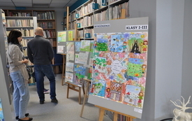 Na planszach między p&oacute;łkami bibliotecznymi prace konkursowe. Obok oglądający prace ludzie.