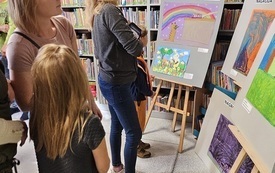 Na planszach między p&oacute;łkami bibliotecznymi prace konkursowe. Obok oglądający prace ludzie.