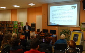 W wnętrzu biblioteki siedzą uczestnicy prelekcji. W tle wyświetlana prezentacja.