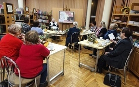 Grupa kobiet siedząca we wnętrzu biblioteki. Na stolikach filiżanki i talerzyki z ciastem.