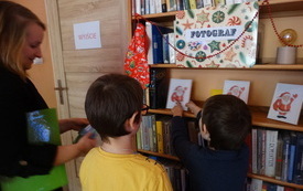 Przy regale bibliotecznym stoi dw&oacute;ch chłopc&oacute;w i kobieta. Na regale Napis fotograf, poniżej trzy obrazki Mikołaja.