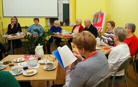 Grupa ludzi siedzi wok&oacute;ł stolik&oacute;w i rozmawia. Na stołach książki, filiżanki. 