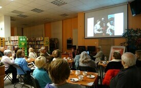 Wnętrze biblioteki, przy stolikach siedzą uczestnicy wykładu. Na ścianie prezentacja. 