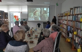 W bibliotece, przy stole siedzą panie seniorki i oglądają wyświetlany film.