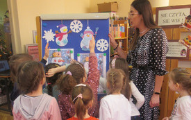 Bibliotekarka stoi wraz z dziećmi przy tablicy, na kt&oacute;rej są papierowe bałwany. 