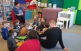 Bibliotekarka trzyma w rękach książkę, na dywanie siedzą dzieci i dorośli. 