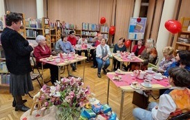 Grupa kobiet siedzi przy stolikach, kobiety zwr&oacute;cone w kierunku prowadzącej spotkanie starszej kobiety.