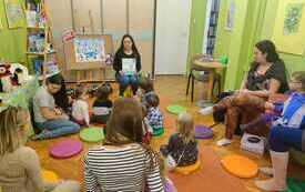 Bibliotekarka siedzi na krzesełku i trzyma książkę, wok&oacute;ł niej siedzą dzieci i rodzice. 