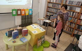 Bibliotekarka z dziewczynką rozwiązują zagadki językowe. Na stoliku stoją słowniki. Na sznurku zawieszone literki. Regały z książkami.