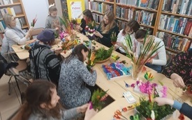 Grono kobiet w bibliotece, siedzących przy stole i tworzących palmy wielkanocne.