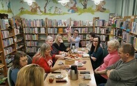 Sala biblioteczna z książkami. Przy stole grupa os&oacute;b dyskutujących o książkach. 