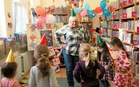 Kobieta wraz z małymi dziewczynkami wśr&oacute;d regał&oacute;w bibliotecznych.