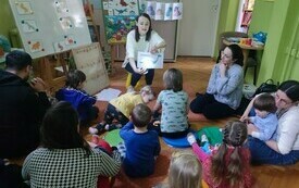 Bibliotekarka pokazuje książkę dzieciom i dorosłym, kt&oacute;rzy siedzą na dywanie.