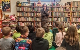 Bibliotekarz prowadzi spotkanie dla dzieci. W tle regały z książkami i prezentacja multimedialna. 