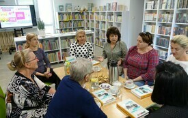 Grupa kobiet siedzi przy stole, na stole książki, filiżanki, w tle regały z książkami. 