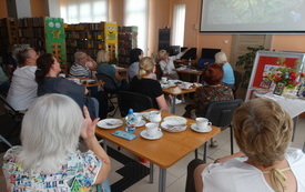 Grupa kobiet siedzi przy stolikach w bibliotece i ogląda prezentację multimedialną, prelegentka wskazuje palcem kwiaty na ekranie