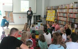 Bibliotekarka pokazuje siedzącym na podłodze dzieciom i ich rodzicom kolorowe obrazki