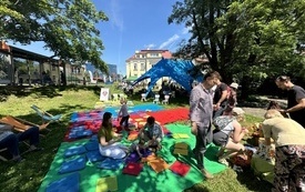 Na trawie rozłożone leżaki, koce i poduszki. Dzieci w raz z rodzicami odpoczywają w cieniu drzew. 