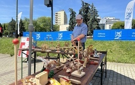 Mężczyzna w okularach i czapce z daszkiem przy stanowisku z drewnianymi zabawkami. 