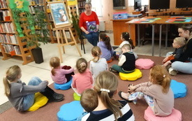 We wnętrzu biblioteki dzieci siedzą na poduszkach i słuchają bibliotekarki, kt&oacute;ra czyta. 