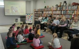 Dzieci zgromadzone w sali bibliotecznej słuchają babć, kt&oacute;re im czytają.