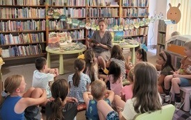 Dzieci pośrodku sali słuchają jak bibliotekarka czyta.