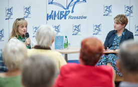 Pisarka po lewej stronie i prowadząca spotkanie po prawej stronie. W tle logo biblioteki z pegazem. 