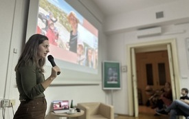 Na pierwszym planie dziewczyna w długich włosach z mikrofonem. W tle widok fotografii na prezentacji. 