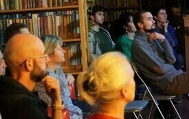 Grupa os&oacute;b siedzących pomiędzy bibliotecznymi regałami i patrzących w tym samym kierunku